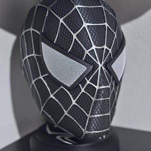 May include: A black Spider-Man mask with silver web detailing and white eye lenses. The mask is displayed on a black stand, showcasing the intricate design and texture of the superhero's iconic headgear.