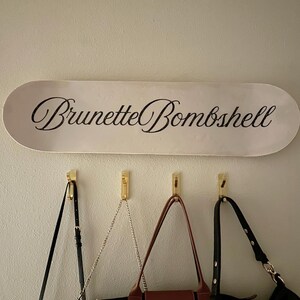 Pode incluir: Uma placa oval branca com as palavras "Brunette Bombshell" em elegante escrita preta est&aacute; pendurada acima de tr&ecirc;s ganchos dourados. Tr&ecirc;s bolsas com al&ccedil;as pretas e marrons pendem dos ganchos, oferecendo uma solu&ccedil;&atilde;o de armazenamento elegante.