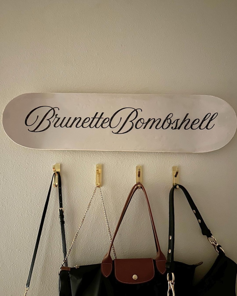 Pode incluir: Uma placa oval branca com as palavras "Brunette Bombshell" em elegante escrita preta est&aacute; pendurada acima de tr&ecirc;s ganchos dourados. Tr&ecirc;s bolsas com al&ccedil;as pretas e marrons pendem dos ganchos, oferecendo uma solu&ccedil;&atilde;o de armazenamento elegante.