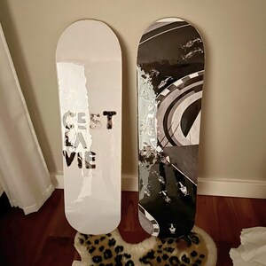Pode incluir: Dois skates encostados a uma parede. Um &eacute; branco com o texto "C'est la vie" em preto. O outro tem um design gr&aacute;fico em preto e branco. As pranchas est&atilde;o sobre um tapete com estampa de leopardo.