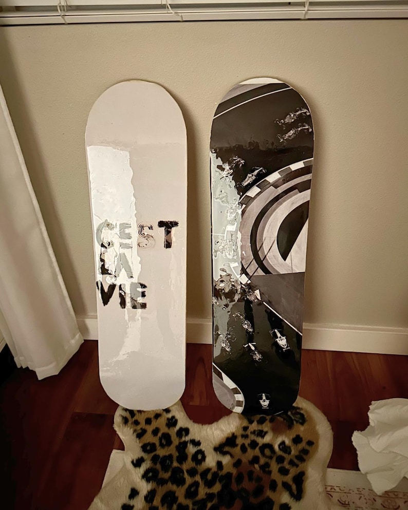 Pode incluir: Dois skates encostados a uma parede. Um &eacute; branco com o texto "C'est la vie" em preto. O outro tem um design gr&aacute;fico em preto e branco. As pranchas est&atilde;o sobre um tapete com estampa de leopardo.