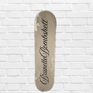 Pode incluir: Um skate com acabamento branco sujo desgastado. As palavras "Brunette Bombshell" est&atilde;o escritas em it&aacute;lico no centro da prancha. A prancha tem uma apar&ecirc;ncia ligeiramente desgastada.