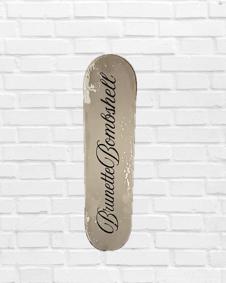 Pode incluir: Um skate com acabamento branco sujo desgastado. As palavras "Brunette Bombshell" est&atilde;o escritas em it&aacute;lico no centro da prancha. A prancha tem uma apar&ecirc;ncia ligeiramente desgastada.
