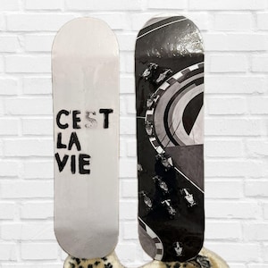 Pode incluir: Dois skates. Um &eacute; branco com o texto "C'est La Vie" em preto. O outro &eacute; preto e branco com um design de carro de corrida. Ambos s&atilde;o exibidos contra uma parede de tijolos brancos.