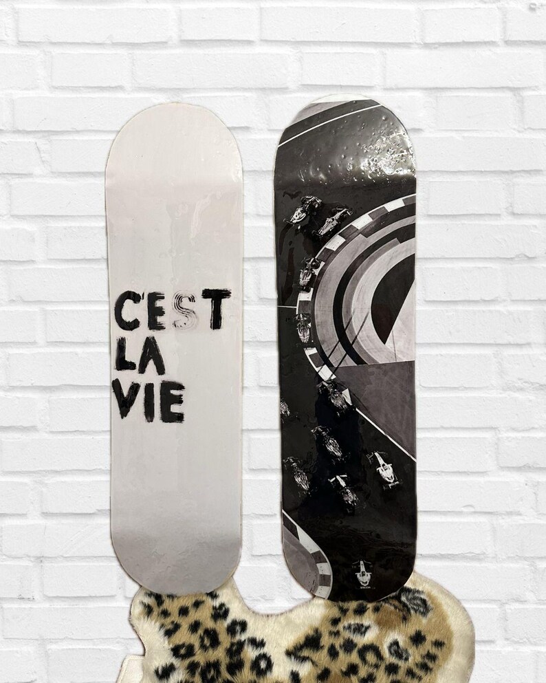 Pode incluir: Dois skates. Um &eacute; branco com o texto "C'est La Vie" em preto. O outro &eacute; preto e branco com um design de carro de corrida. Ambos s&atilde;o exibidos contra uma parede de tijolos brancos.