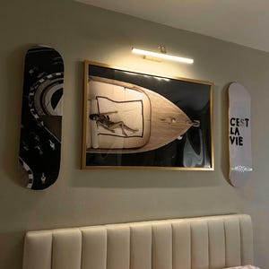 Pode incluir: Uma parede de quarto apresenta uma obra de arte emoldurada com uma pessoa em um barco, ladeada por dois skates. Um skate tem um design em preto e branco, o outro diz "C'est La Vie". Uma lumin&aacute;ria est&aacute; acima da obra de arte.