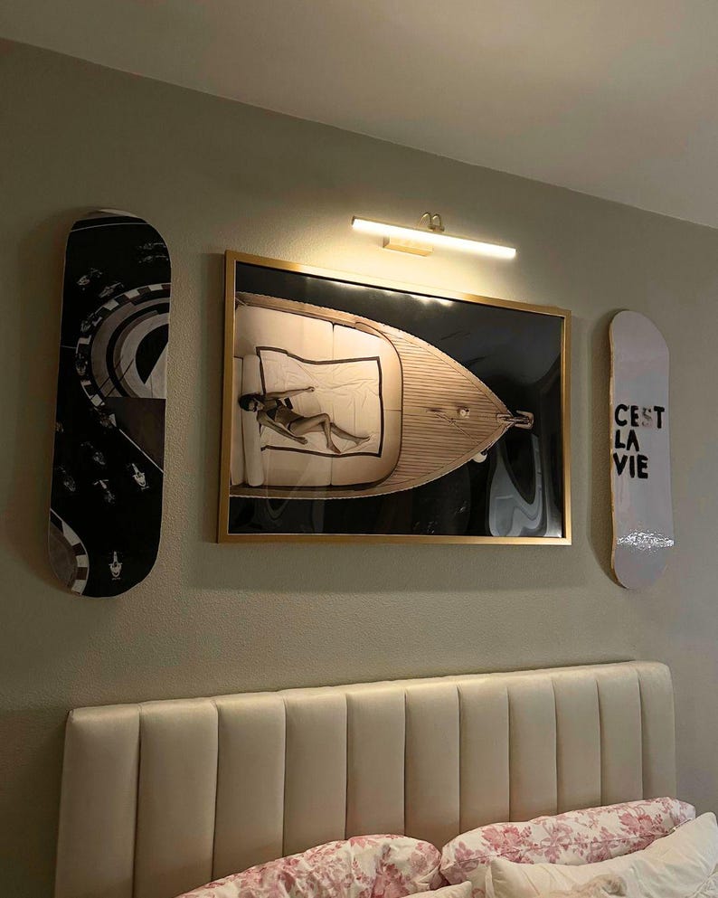 Pode incluir: Uma parede de quarto apresenta uma obra de arte emoldurada com uma pessoa em um barco, ladeada por dois skates. Um skate tem um design em preto e branco, o outro diz "C'est La Vie". Uma lumin&aacute;ria est&aacute; acima da obra de arte.