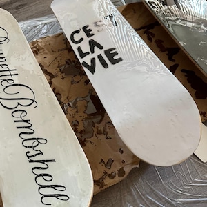 Pode incluir: Tr&ecirc;s skates com superf&iacute;cies brancas. Uma prancha tem a frase "C'est La Vie" em preto, outra a palavra "Brunette Bombshell" em it&aacute;lico e a terceira um design preto e prateado. As pranchas est&atilde;o sobre uma superf&iacute;cie castanha.