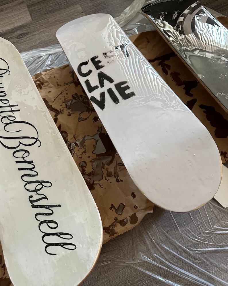 Pode incluir: Tr&ecirc;s skates com superf&iacute;cies brancas. Uma prancha tem a frase "C'est La Vie" em preto, outra a palavra "Brunette Bombshell" em it&aacute;lico e a terceira um design preto e prateado. As pranchas est&atilde;o sobre uma superf&iacute;cie castanha.