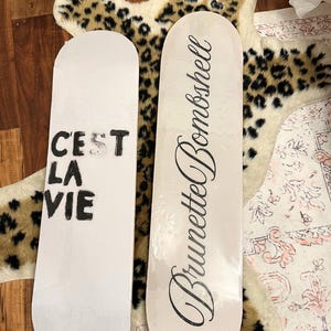 Pode incluir: Duas pranchas de skate brancas. Uma tem o texto "C'EST LA VIE" em preto, a outra l&ecirc; "Brunette Bombshell" em it&aacute;lico. As pranchas est&atilde;o sobre um tapete com estampa de leopardo.