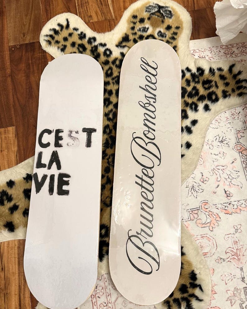 Pode incluir: Duas pranchas de skate brancas. Uma tem o texto "C'EST LA VIE" em preto, a outra l&ecirc; "Brunette Bombshell" em it&aacute;lico. As pranchas est&atilde;o sobre um tapete com estampa de leopardo.