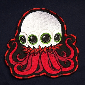 May include: Embroidered patch featuring a cartoon octopus-like creature with a white head, four green eyes, and red tentacles. The patch has a black border with red accents and is set against a dark blue background.