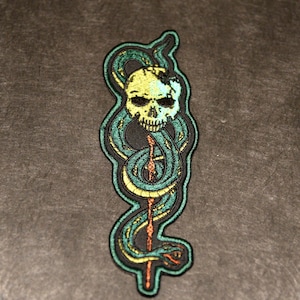 May include: Embroidered patch featuring a skull with a snake wrapped around it. The skull is yellow with a crack, and the snake is green and yellow. The patch has a black border and is on a dark gray background.