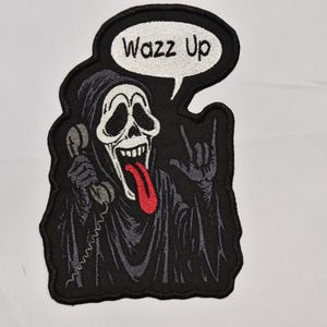 May include: Embroidered patch featuring the Ghostface character from the Scream movies. The design shows Ghostface holding a phone, with a red tongue sticking out, and the text "Wazz Up" in a speech bubble. The patch is black, white, and gray.