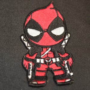 May include: Embroidered patch featuring a cartoon version of Deadpool. The patch is primarily red and black, with white accents on the eyes and belt. The character is depicted with two swords on his back.