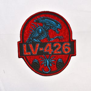 May include: Embroidered patch featuring a blue alien creature design on a red background, with the text "LV-426" in orange. The patch also includes a stylized image of eggs and a smaller alien. The patch is circular with a black border.