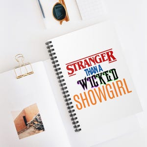 Puede incluir: Cuaderno blanco con espiral con el texto "STRANGER THAN A WICKED SHOWGIRL" en rojo, negro, verde, morado y naranja. El cuaderno está abierto, revelando una página en blanco y una foto. Gafas de sol y un clip también son visibles.