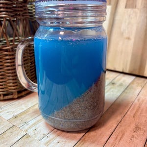 May include: A clear glass mason jar with a handle, filled with blue liquid and sand. The jar has a clear lid and is sitting on a wooden surface. A wicker basket is in the background.