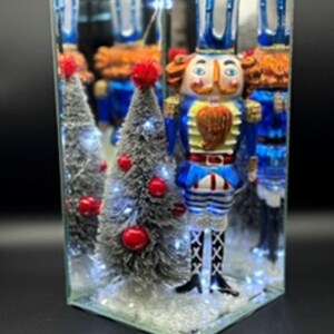 May include: A glass nutcracker figurine in a blue and white uniform with a gold beard, standing next to a small, flocked Christmas tree decorated with red ornaments and white LED lights, all enclosed within a mirrored, triangular display case.