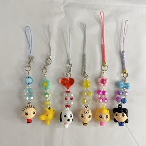Op de afbeelding: Zes kralen telefoonbedels met stripfiguren. Elke bedel heeft een ander ontwerp, waaronder een jongen, een gele hond, een witte hond, een jongen, een meisje en een meisje. De bedels hebben verschillende kleuren kralen en bijpassende gekleurde koorden.