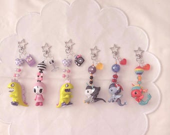 May include: Six decorative keychains featuring various miniature figures including yellow dinosaurs, a pink skull character, and mythical creatures. Each keychain has a silver star-shaped clasp, colorful plastic beads, and small charms.