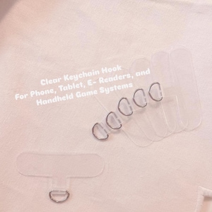 Op de afbeelding: Transparante sleutelhaken met zilverkleurige ringen. De tekst op de afbeelding luidt: Clear Keychain Hook For Phone, Tablet, E-Readers, and Handheld Game Systems. De haken zijn ontworpen voor verschillende apparaten.