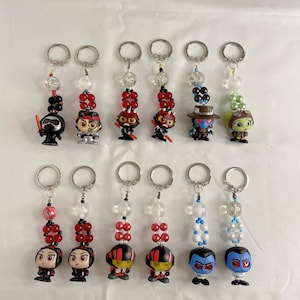 Puede incluir: Colección de llaveros de personajes con detalles de cuentas. Cada llavero presenta una pequeña figura unida a un llavero plateado. Las cuentas son transparentes, rojas y blancas. Las figuras representan varios personajes.