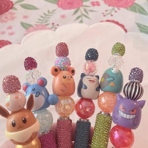 May include: A selection of decorative pens with character toppers. Each pen has a jeweled barrel and is adorned with a different character, such as a brown Eevee, blue Azurill, orange Teddiursa, and purple Gengar. The pens are displayed against a floral backdrop.