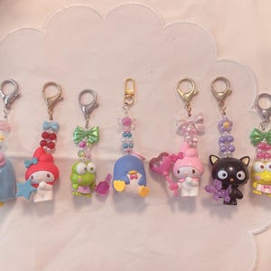 Puede incluir: Una colección de llaveros de personajes coloridos. Cada llavero presenta un personaje de dibujos animados diferente, como un pingüino, una rana, un gato y otros. Los llaveros están sujetos a cierres plateados y adornados con detalles de cuentas.