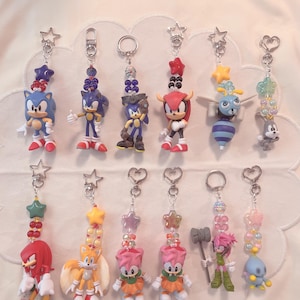 May include: A selection of character keyrings featuring Sonic the Hedgehog and friends. Each keyring has a star or heart-shaped charm, colourful beads, and a silver-tone clasp. The figurines include Sonic, Tails, Amy Rose, Knuckles, and others, in various poses.