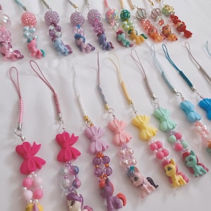 Puede incluir: Una colección de colgantes para teléfono coloridos con pequeñas figuras de pony, lazos y detalles de cuentas. Los colgantes tienen varios esquemas de color, incluyendo rosa, morado, amarillo y azul, con herrajes plateados y cordones a juego.