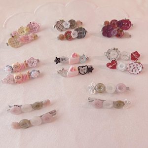 May include: A collection of metal hair clips decorated with various buttons, charms, and miniature figures. The designs include cats, cakes, hearts, flowers, and plaid patterns in shades of pink, white, green, and red, arranged in pairs.