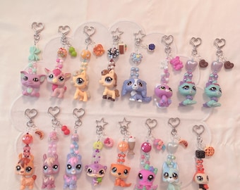 May include: A collection of colorful keychains featuring various animal figurines. Each keychain has a silver-tone heart-shaped clasp and is adorned with beads and charms in shades of pink, blue, orange, and green. The figurines include cats, dogs, and other creatures.