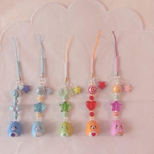 May include: Five phone charms featuring small bear figurines in blue, light blue, green, orange, and pink. Each charm includes colourful plastic beads, star-shaped accents, and a spiral candy-like bead attached to a thin nylon cord loop.