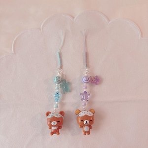 May include: Two phone charms featuring brown bear figures wearing sleep masks. The left charm has blue beads, a fish, and a turtle. The right charm has purple beads, a swirl, and a star. Both charms hang from thin, coloured cords.
