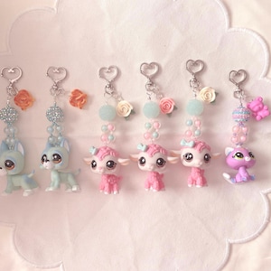 May include: Six decorative keychains featuring small animal figurines. Two light blue dogs with orange flower charms, three pink lambs with rose charms, and one purple cat with a pink bear charm. Each includes heart-shaped clasps and beads.