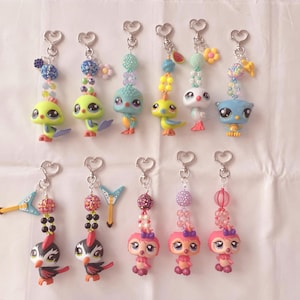 May include: A collection of colorful keychains featuring various animal figurines. Each keychain has a silver heart-shaped clasp and is adorned with beads in shades of blue, pink, and yellow. The figurines include birds and other creatures.