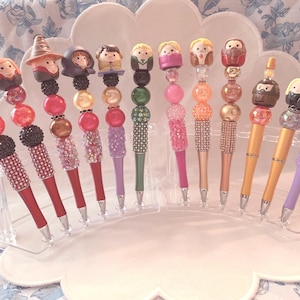 Op de afbeelding: Een verzameling decoratieve pennen met karakterthema-toppers. De pennen hebben verschillende kleuren, waaronder rood, roze, groen en paars, met met strass bezette barrels en kralenaccenten. De toppers tonen stripfiguren.