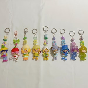 Puede incluir: Una colección de ocho llaveros de personajes coloridos. Cada llavero presenta una pequeña figura con un atuendo único y una cadena de cuentas. Las figuras son de varios colores, incluyendo amarillo, rosa, azul y verde, con llaveros plateados.