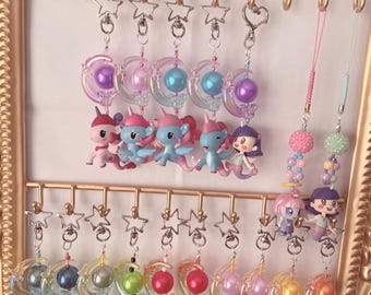 May include: A collection of colorful keychains featuring various cartoon characters, including unicorns and mermaids. Each keychain has a star-shaped clasp and a decorative charm. The keychains are displayed on a gold-colored rack.