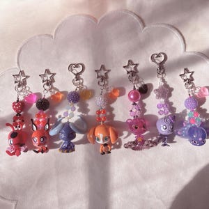 May include: Seven decorative keychains featuring small cartoon character figurines attached to silver-toned star or heart-shaped clasps. Each keychain includes a variety of colorful plastic beads, including textured spheres and small heart charms.