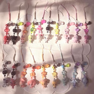 May include: A collection of twenty phone charms featuring small character figurines, colorful plastic beads, and star or heart-shaped accents. Each charm is attached to a thin cord loop, showcasing a variety of pastel and vibrant color palettes.