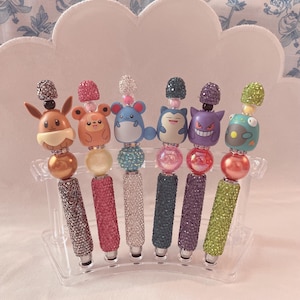 Könnte beinhalten: Sechs dekorative Stifte mit Charakter-Designs. Jeder Stift hat einen anderen Charakter auf der Oberseite und einen mit Juwelen besetzten Schaft in verschiedenen Farben. Die Stifte werden in einem transparenten Kunststoffhalter präsentiert.