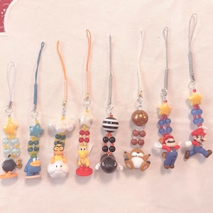 May include: A collection of eight colorful phone charms featuring various cartoon characters and shapes. Each charm has a beaded strand with a star or other shape at the top and a small figurine at the bottom. The charms include stars, clouds, and characters.