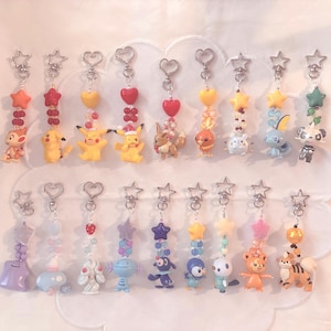 May include: A collection of eighteen keychains featuring various small plastic character figures attached to metal star-shaped clasps. Each keychain includes a vertical arrangement of colorful beads, stars, and heart-shaped charms in diverse hues.