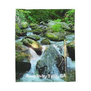 May include: A printed throw blanket featuring a scenic view of Anna Ruby Falls, GA. The image shows a flowing stream cascading over moss-covered rocks, surrounded by lush green foliage. The text "Anna Ruby Falls, GA" is visible.