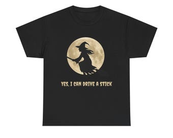 May include: A black short-sleeved t-shirt featuring a graphic of a witch flying on a broomstick in front of a large, pale yellow full moon. Below the graphic, the text reads, "YES, I CAN DRIVE A STICK" in a light-colored, distressed font.