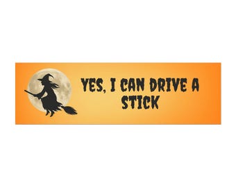 May include: A rectangular orange bumper sticker featuring a black silhouette of a witch flying on a broomstick in front of a full moon. The text in black, stylized font reads: YES, I CAN DRIVE A STICK. The design is centered on a solid orange background.