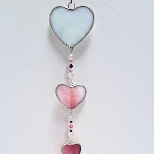 May include: A hanging heart-shaped suncatcher with a silver-colored metal frame. The suncatcher features three hearts in shades of pink and a clear heart at the bottom, connected by a string of beads. The top heart is a pale blue color.