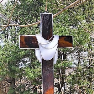 May include: A stained glass cross with a white, translucent, fabric-like design in the center. The cross is made of dark brown glass with a reddish-brown hue.